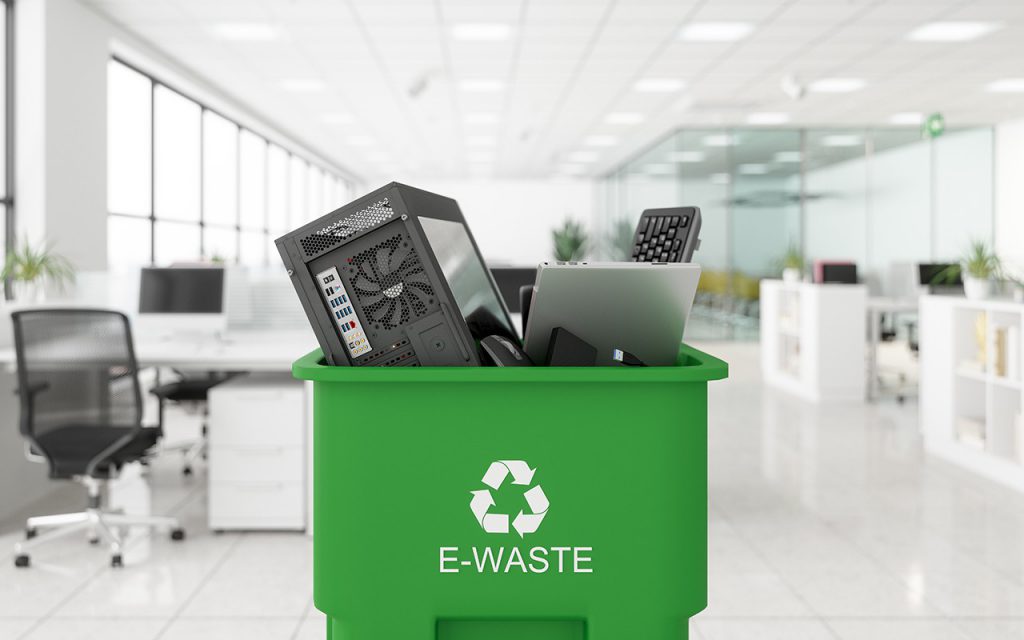 ถังขยะสีเขียวติดคำว่า E-WASTE ข้างในมีเคสคอมพิวเตอร์ คีย์บอร์ด ซึ่งเป็นขยะอิเล็กทรอนิกส์