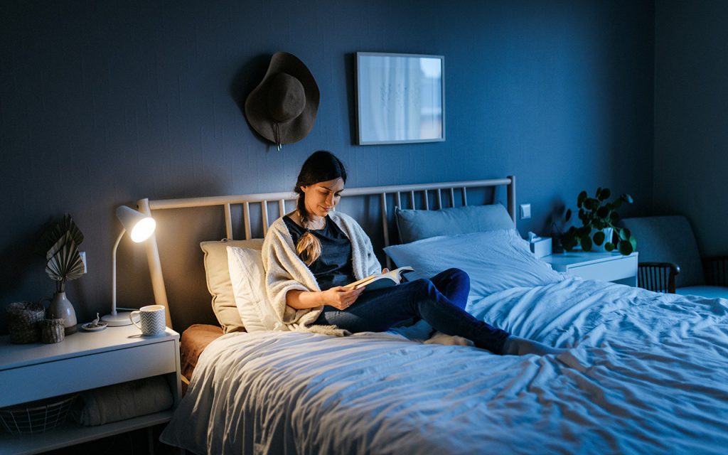 หญิงสาวอ่านหนังสือบนเตียง มีโคมไฟเปิดอยู่ขัางหัวเตียง