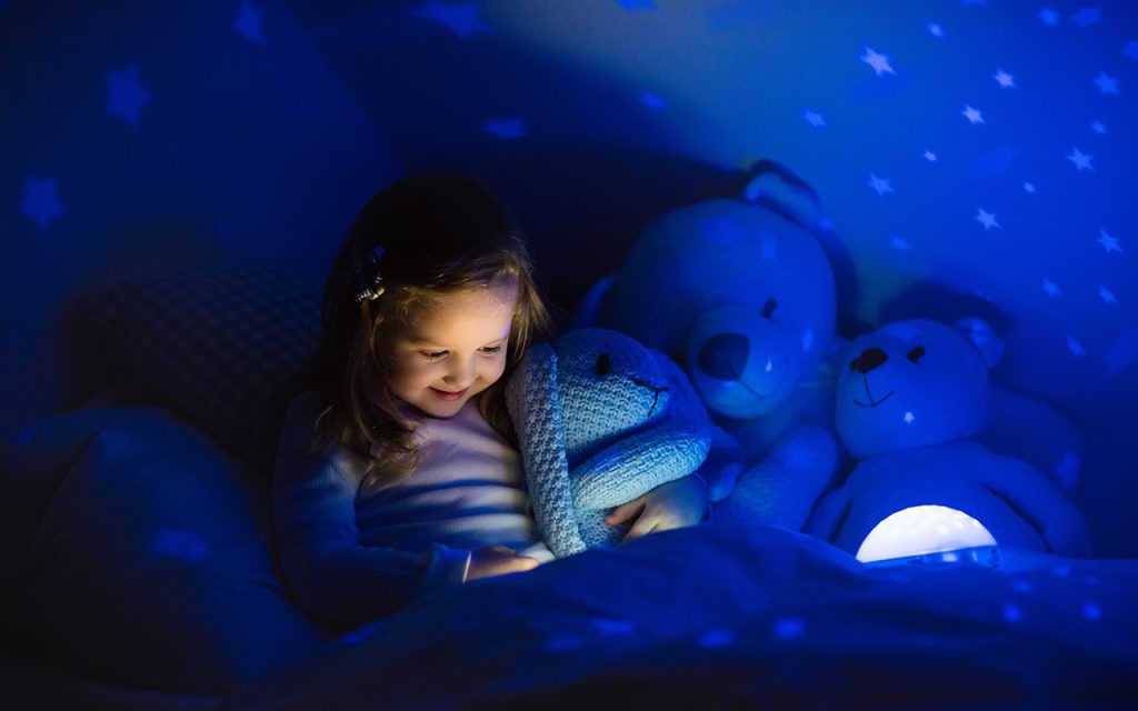 เด็กผู้หญิงนั่งเอนตัวกอดตุ๊กตากระต่ายหูยาวไว้ในแขนขณะนอนดูบางอย่างอยู่บนเตียง